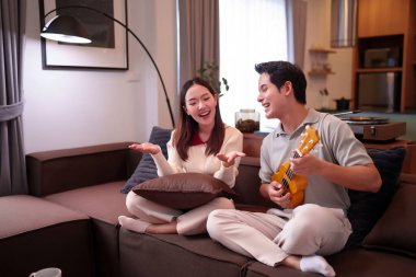 Asyalı yetişkin bir çift neşeli bir atmosferle evdeki kanepede ukulele çalıyor. Asyalı bir kadın gülümser ve tatlı bir melodi boyunca şarkı söyler..