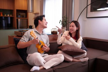 Neşeli bir Asyalı çift ukulele çalmaktan ve kanepede birlikte gülmekten zevk alır. Evde eğlenen yetişkin kadın ve erkek anları yaratırlar..