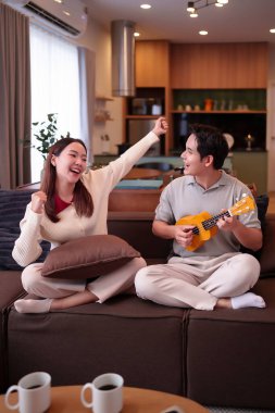 Asyalı bir çiftin sıcak oturma odalarında hoş bir sahnesi. Genç adam bir gülümsemeyle ukulele çalarken, genç kadın sevinçle kutluyor, paylaşılan müzik ve mutluluğun neşeli bir anını sergiliyor..