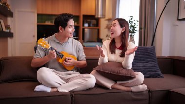 Genç bir Asyalı çiftin keyifli zamanlarını yakalamak erkeğin ukulele çalarken kadın mutlu bir şekilde dinlerken sıcak evlerinde güzel anılar yaratıyor..