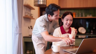 Genç Asyalı çift evde dizüstü bilgisayarla çalışırken gülümsüyor. Mutlu bir ilişkinin tadını çıkaran çift, serbest ve teknolojik yaşam tarzı için kahveyle rahatlıyor..
