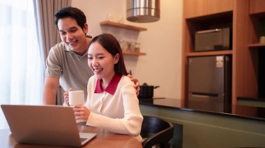 Asyalı bir çiftin evde kaliteli zaman geçirdiği o güzel anı yakalayın. Asyalı bir adam dizüstü bilgisayar kullanırken Asyalı bir kadın elinde bardak tutuyor, hem gülüyor hem de gülümsüyor ve mutluluklarını gösteriyor.