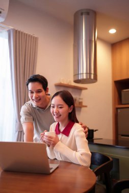 Genç bir Asyalı çift evde dizüstü bilgisayar kullanırken gülümserken kadın elinde bir fincan kahve tutuyor. Onlar mutlular ve evlerinde çevrimiçi olarak vakit geçiriyorlar..