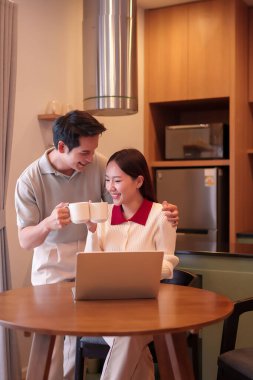 Neşeli bir Asyalı çift evde elinde kahve fincanlarıyla dizüstü bilgisayarla çalışırken gülümsüyor. Modern yaşam tarzını, tatlı anı ve sevgi dolu bir ilişkiyi yakalar..