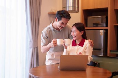 Asyalı bir çiftin mutluluğunu kahve içerek ve dizüstü bilgisayarla mutfaklarında yakalamak. Yetişkin kadın ve erkek için rahat bir bağlantı, iş ve boş zaman sahnesi.