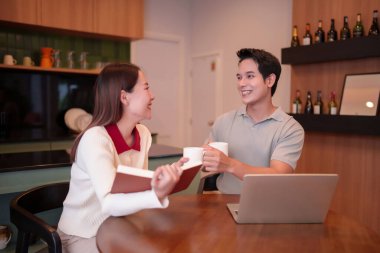 Asyalı bir adamın ev ofisinde kahve kupası sunarken elinde kitap tutan Asyalı genç bir kadının çekici görüntüsü. Dizüstü bilgisayarın üstünde şarap şişeleri sergileniyor..