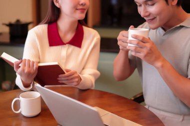 Evde eğlenen genç bir Asyalı çiftin görüntüsü. Asyalı bir adam kahve içerken Asyalı bir kadın da tahta bir masada dizüstü bilgisayarın yanında kitap okuyor. Birliktelik kavramı