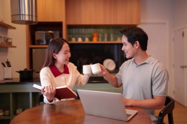 Mutlu genç Asyalı çift evlerinin mutfağında kahve fincanları kızartıyorlar, masada dizüstü bilgisayar ve dizüstü bilgisayarlarıyla keyifli bir anın tadını çıkarıyorlar. Boş yaşam tarzı konsepti.