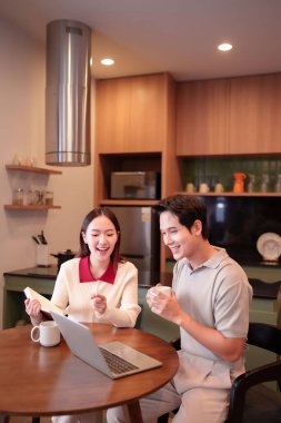 Yetişkin bir Asyalı elinde kahve fincanı tutarken genç bir Asyalı kadın kitap okuyor. Modern bir mutfakta dizüstü bilgisayarda birlikte çalışıyorlar. Fikirlerini paylaşmaktan mutluluk duyarlar..