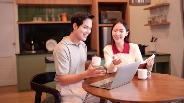 Gülümseyen Asyalı yetişkin bir çift evde dizüstü bilgisayarda bir şey tartışırken kahve içiyor. Kadın elinde bir kitap tutuyor, modern bir çalışma alanında işbirlikçi ve sıradan bir yaşam tarzını teşvik ediyor..
