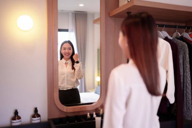 Bu resim, kendine güvenen Asyalı bir kadının gülümseyişini gösteriyor, yatak odasındaki ayna yansımasına bakıyor. Çok şık giyiniyor, iş kıyafeti giyiyor ve moda dolabının yanında hazırlanıyor..