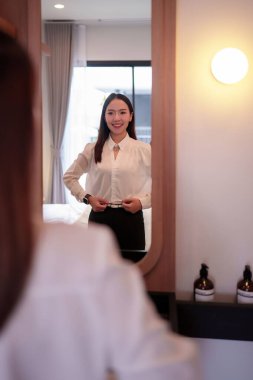 Aynaya yansıyan Asyalı bir kadın kemerini ayarlıyor ve şık kıyafetlerini tamamlıyor. Bir otel banyosunda hazırlanırken, bu görüntü onun tarzına ve detaylarına olan ilgisini çeker..