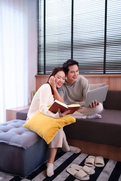Mutlu Asyalı çift evdeki kanepede birlikte dinleniyor. Yetişkin adam dizüstü bilgisayar kullanıyor ve kadın kitap okuyor. Yastık ve pencereyle güzel vakit geçiriyor..