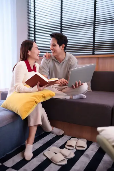 Bu resimde Asyalı yetişkin bir çiftin evde dinlendiği görülüyor. Kadın dizüstü bilgisayarını kullanırken elinde bir kitap tutuyor. Gülümsüyor.
