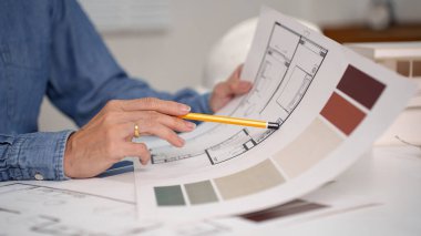 Yaşlı bir yetişkinin eli, bir iç tasarım projesini simgeleyen plan ve renk örneklerinin üzerinde bir kalem tutuyor. Bu detaylı sahnede mimari tasarım ve ev yenileme fikirlerini araştırın. Yaşlı yetişkin el inte için renk paletlerini dikkatlice seçer