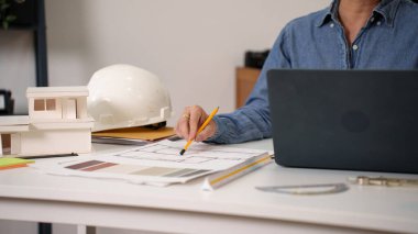 Yetişkin Asyalı mühendis el, kalemle plan çiziyor. Dizüstü bilgisayarı ve beyaz şapkası olan bir masa üzerinde ev tasarımı yapmak. Ev tasarımı için mimari el çizimi planı, güvenlik ve işlevselliğin sağlanması için hayati bir süreç.