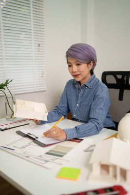 Asyalı yaşlı kadın mimar, masa üzerinde tasarımı olan ev modeli projesi tasarlıyor. O, son tasarım fikirleri üzerinde çalışıyor. Bu Asyalı yaşlı kadın mimar ev modelini dizayn edişinin bir kanıtıdır..