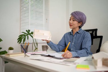 Asyalı yaşlı bir mimar, ofis masasındaki planları incelerken titizlikle bir bina modeli tasarlıyor. Asyalı yaşlı kadın mimar, mimari tasarım ve geliştirmede profesyonel uzmanlık sergiliyor.