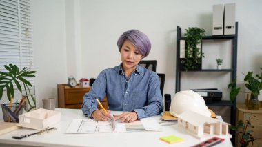 Bir Asyalı Yaşlı Kadın Mimar ev tasarımı için bir plan çiziyor. Asyalı Yaşlı Kadın Mimarı, konut projesinin planları üzerinde çalışıyor..