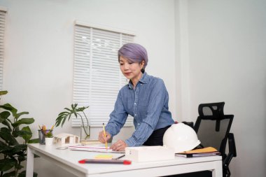 Ofisinde titizlikle bina planları tasarlayan yaşlı bir Asyalı kadın mimar imajımızı keşfedin. Bu resim, yeni bir inşaat projesinin mimari taslağını hazırlamak için bir mimar kadını ustalıkla kalem ve cetvel kullanarak sergiliyor.