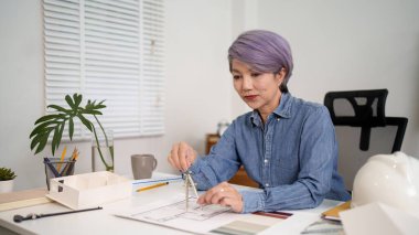 Yaşlı Asyalı kadın mimar titizlikle mimari tasarım projesini çiziyor. Yapı inşaatı için pusulası olan taslak planına odaklanıyor ve çalışmasında kesinlik sağlıyor. Yaşlı Asyalı kadın..