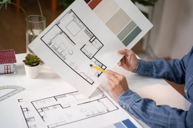 Asyalı yetişkin eller, iç dizayn planının üzerinde bir kalemle işaret ediyorlar. Kat planının gözden geçirilmesi, ev mimarisi projesinin bir parçası. Ayrıntılı olarak gösterilen yaşam alanlarının yaratıcı yenilenmesi. Bir iç tasarım planı keşfet