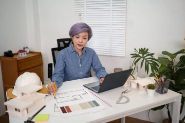 Asyalı yaşlı bir mimar kadın dizüstü bilgisayarı üzerinde ev planı tasarımlarıyla çalışıyor. O bir profesyonel, bir ev yenileme projesi planlıyor. Bu resimde gösterilen mimarlık tasarım uzmanlığı, özverisini vurguluyor.