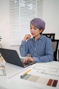 Asyalı yaşlı bir kadın, mimarlık projesini tasarlamak için evden dizüstü bilgisayar üzerinde çalışıyor. Asyalı yaşlı bir kadın ev mimarı projesi tasarlıyor.