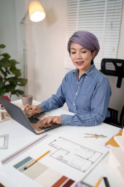 Yaşlı Asyalı kadın mimar bir bina planı tasarlamak için dizüstü bilgisayar üzerinde çalışıyor. Yaşlı Asyalı kadınlar ev içi ve mimari projeler tasarlıyorlar. Kat planlarını mavi gömlekle hazırlıyorlar..