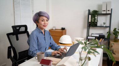 Yaşlı bir Asyalı kadın mimar, müşterileri için bir ev planı hazırlamak için dizüstü bilgisayarını kullanıyor. Kendisi, iş yerinde yazılım araçlarıyla bina planları yapan yaşlı bir Asyalı kadın mimar..