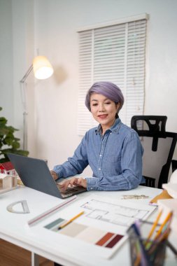 Yaşlı bir Asyalı mimarın dizüstü bilgisayarını kullanarak özenle bir yapı planı üzerinde çalıştığı profesyonel mimari tasarım projesi. Bu Asyalı kadın mimar, parlak bir evde müşteri projesi için mimari tasarım ve detaylara odaklanıyor.