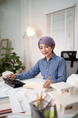 Yetişkin Asyalı kadın mimar, tablet kullanarak ve model evle birlikte bir deftere yazı yazarak iç tasarım için bir ev planı hazırlıyor. Kıdemli işletme sahibi tasarım projesi ve tasarım planı.