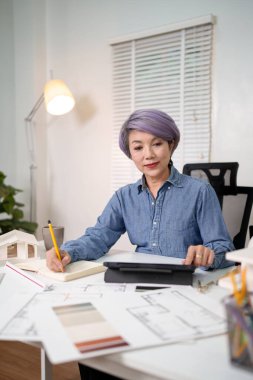 Yaşlı bir Asyalı mimar ofisinde bir ev planı tasarlıyor. Mimari tasarım projesi dijital tablet kullanır ve not alır. Bu mimar ev planı planını ustalıkla tasarlıyor..