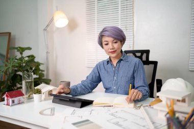 Yaşlı Asyalı kadın mimar tablet kullanarak bir ev planı tasarlıyor. Üst düzey bir mimar masasında çalışıyor, ev tasarımı fikirlerini analiz ediyor. Asyalı kadın mimar odaklanmış ve kendini işine adamış..