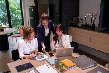 Modern bir ofiste işbirliği yapan üç yetişkin Asyalı kadın. İş stratejilerini tartışmak, verileri analiz etmek ve profesyonel bir çalışma ortamında dizüstü bilgisayarlar ve hesap makineleriyle plan yapmak..