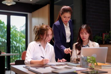 İş toplantısında, profesyonel kıyafetli genç Asyalı kadınlar, başlangıç büyümesi için takım çalışması koçluğu ve profesyonel gelişimi vurgulayan bir ofiste iş birliği yapıyorlar.