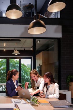 Üç genç Asyalı kadın modern bir ofis alanında iş toplantısı yapıyorlar. Bir dizüstü bilgisayar kullanarak ve kurumsal bir ortamda fikirleri tartışarak bir projede işbirliği yapıyorlar. Bu bir takım çalışması göstergesidir..