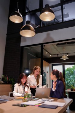 Üç genç Asyalı kadın modern bir ofiste üretken bir takım toplantısı yapıyorlar. İş stratejilerini tartışıp bir proje üzerinde birlikte çalışıyorlar. İşbirliği ve takım çalışması.