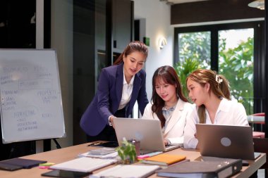 Çeşitli yaşlardan üç Asyalı kadın, bir proje tanıtımı sırasında modern ofis ortamında dizüstü bilgisayar kullanarak bir iş planı üzerinde işbirliği yapıyorlar.