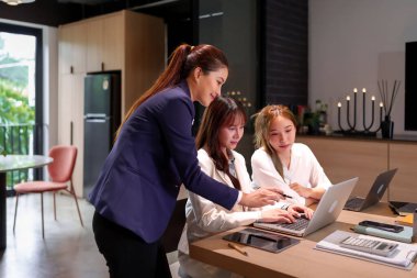 Modern bir ofis ortamında üç Asyalı kadın işbirliği yapıyor. Bir kadın dizüstü bilgisayarda çalışan iki genç profesyonele akıl hocalığı yapıyor. Bu görüntü, kadın takım çalışmasının güçlendirilmesini ve destekleyici bir çalışma ortamını temsil ediyor..