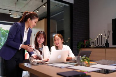 Modern bir ofiste işbirliği yapan üç Asyalı kadın. Dizüstü bilgisayar kullanıyorlar ve bir projeyi tartışıyorlar. Bu resim takım çalışmasını, iş başarısını ve iş yerinde teknolojinin kullanımını gösteriyor..