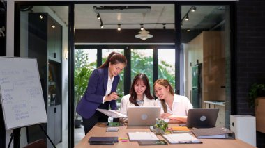 Modern bir ofiste üç Asyalı kadın bir iş planı üzerinde çalışıyor. Bir proje üzerinde işbirliği yapıyorlar, laptoptaki verileri analiz ediyorlar ve bir pazarlama stratejisi yaratıyorlar. Atmosfer pozitif ve işbirlikçi, takım çalışmasını gösteriyor..
