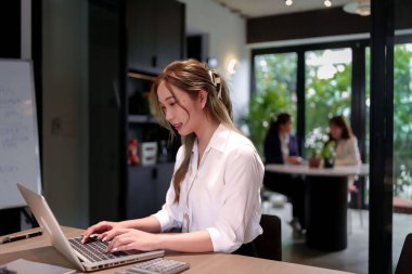 Bu resim, parlak, modern bir ofiste dizüstü bilgisayarıyla çalışan genç bir Asyalı kadını tasvir ediyor. Üretken bir şirket atmosferine katkıda bulunan görevlerine odaklanmış durumda. Verimliliği temsil ediyor..