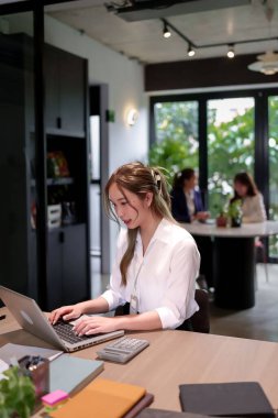 Genç Asyalı kadınlar, iş başarısını artırmak için hesap makineleri ve not defterleriyle modern ofislerdeki dizüstü bilgisayarlarda ve masadaki diğer takım üyeleriyle birlikte çalışıyorlar.