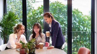 Üç Asyalı kadın, modern ve parlak bir ofiste iş birliği yapıyorlar. Tablete bakıp kahve içiyorlar. Arkadaşça iş yerlerinin tadını çıkarıyorlar.