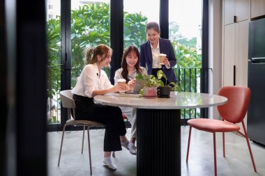 Modern bir çalışma alanında kahve molası veren üç Asyalı kadın dizüstü bilgisayar kullanarak takım çalışması ve iletişim kuruyor.