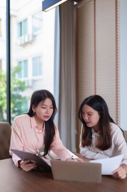 İki genç Asyalı kadın, parlak ve modern bir ofiste bir iş projesinde işbirliği yapıyorlar. Veriler analiz ediliyor, stratejiler tartışılıyor, takım çalışması için dizüstü bilgisayar ve tablet kullanılıyor ve başarıya ulaşılıyor.