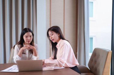 Görüntüde iki yetişkin Asyalı kadın bir iş toplantısında dizüstü bilgisayardan mali raporları tartışıyor. İş yerinde takım çalışması, veri analizi ve proje stratejisini göstermek için ideal.