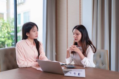 Finansal verileri analiz eden iki yetişkin Asyalı kadının bir iş toplantısında görüntüsü. Modern ofis ortamında iş stratejilerini tartışmak ve raporları gözden geçirmek için bir dizüstü bilgisayar ve hesap makinesi kullanıyorlar..