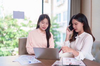 Bu görüntüde iki yetişkin Asyalı kadın bir iş ortamında dizüstü bilgisayardaki verileri analiz ederken, bir tanesi de telefon görüşmesi yapıyor. Bir masada stratejilerini tartışıyor ve büyüme için mali raporlar üzerinde işbirliği yapıyorlar..